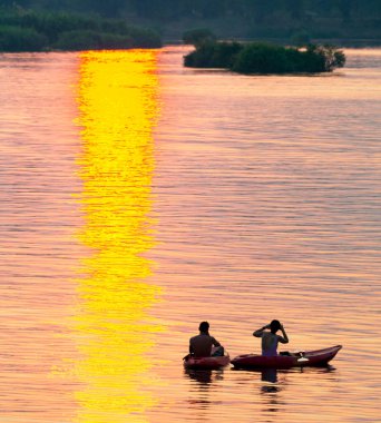 Kayaklardaki iki insan figürünün siluetleri Mekong 'un sakin ve huzurlu sularında yüzüyor. Güney Laos' taki güneşten gelen, suya, batan güneşe yansıyan altın ışık ışınları aracılığıyla..