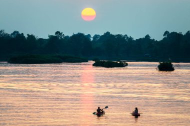 Kayaklardaki iki insan figürünün siluetleri Mekong 'un sakin ve huzurlu sularında yüzüyor. Güney Laos' taki güneşten gelen, suya, batan güneşe yansıyan altın ışık ışınları aracılığıyla..