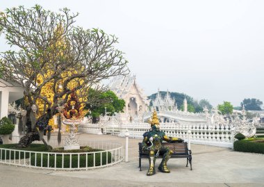 Chiang Rai, Tayland-29 Mart 2023: Turistleri fotoğraf çekmeye ikna etmek için usta sanatçı Chalermchai Kositpipat tarafından tasarlanan ünlü tapınağın ve büyük sanat eserlerinin giriş kapısının yanındaki bankta oturuyor..
