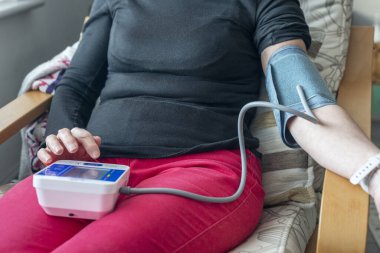 Sağlık bilinci yerinde bir kadın, koltuğunda, evinde, tansiyon monitörüyle, sağlık rejiminin bir parçası olan genel fiziksel durumunu kontrol ederek sağlıklı bir yaşam tarzını sürdürüyor..