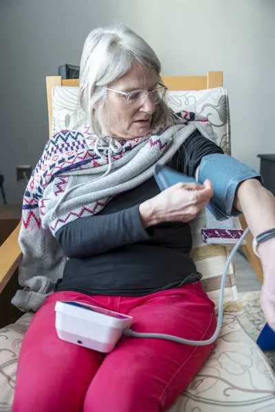 stock image A health conscious female,sits in an armchair,in her house,using a blood pressure monitor to self-check her general physical condition,part of her health regime,to maintain a healthy lifestyle.