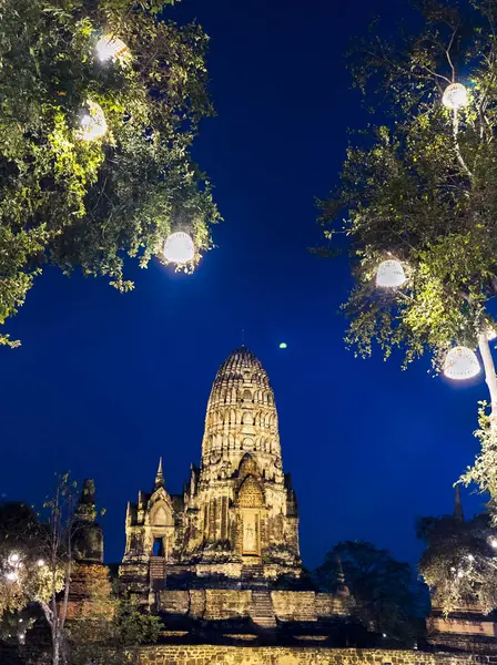ランプは夕暮れの木から吊り下げられ 寺院は明確な暗い青い空と月に対して洪水の照明によって点灯されます — ストック写真