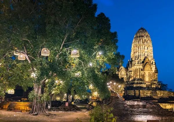 ランプは夕暮れの木から吊り下げられ 寺院は明確な暗い青い空と月に対して洪水の照明によって点灯されます — ストック写真