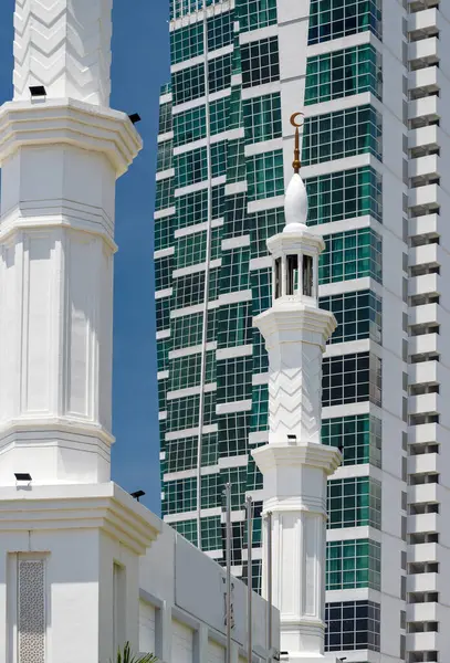 stock image A tall white minarete,In brilliant sunlight,against clear blue sky,near the seafront in Georgetown,Penang's capital,standing close to a high end skyscraper.