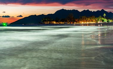 Popüler turistik sahil beldesi, gün batımından sonra, dingin, ruhani bir yer, altından sonra ışıltı, pembe gökyüzü, suya yansıyan ışıklar, sakin denizin hareket bulanıklığı, ve ötesindeki dağların silueti.