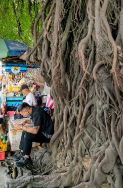 Manila-January 13 2023:People selling small food and drinks at streetside stalls,sit next to wild tree roots,creeping over the wall behind. clipart