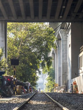 Bangkok, Tayland-07 Nisan 2024-Şehir demiryolu rayları güneşli bir öğleden sonra, büyük bir beton üstten tren yolunun gölgesinde, geçmişteki gecekondu evlerinden geçiyor..