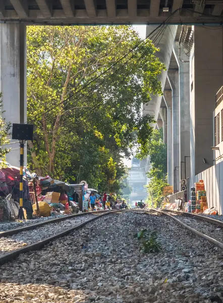 Bangkok, Tayland-07 Nisan 2024-Şehir demiryolu rayları güneşli bir öğleden sonra, büyük bir beton üstten tren yolunun gölgesinde, geçmişteki gecekondu evlerinden geçiyor..