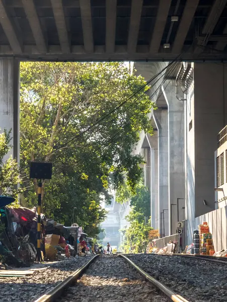 Bangkok, Tayland-07 Nisan 2024-Şehir demiryolu rayları güneşli bir öğleden sonra, büyük bir beton üstten tren yolunun gölgesinde, geçmişteki gecekondu evlerinden geçiyor..