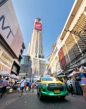 Tayland-Nisan 07-2024-Bir araba, Baiyoke Sky Hotel Kuleleri 'nin ötesindeki güneş ışığına doğru öğleden sonra pazarında alışverişçilerin yanından yavaşça geçiyor..