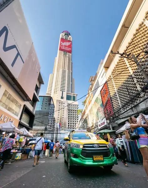 Tayland-Nisan 07-2024-Bir araba, Baiyoke Sky Hotel Kuleleri 'nin ötesindeki güneş ışığına doğru öğleden sonra pazarında alışverişçilerin yanından yavaşça geçiyor..