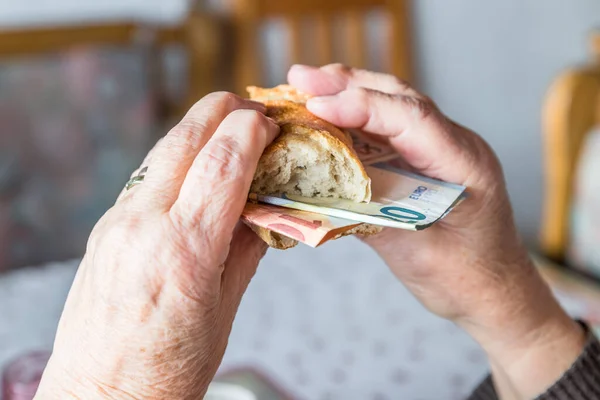 Old woman granny pensioner sits at set breakfast table eating a roll covered with euro banknotes , Germany