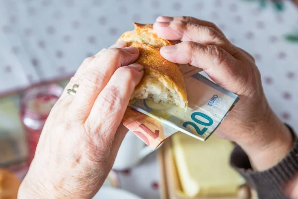 Old woman granny pensioner sits at set breakfast table eating a roll covered with euro banknotes , Germany