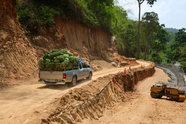A pickup truck transports a truck full of fresh bananas. road construction is going on. A country road route through a valley along a river where the road is damaged during the rainy season.