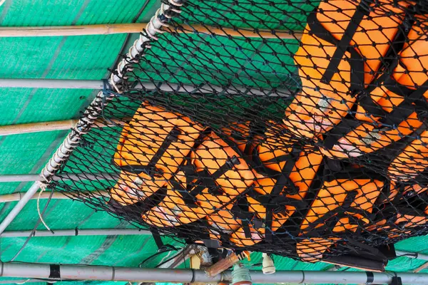 Several orange life jackets are in a net under the canopy of a tour boat. that is about to play into the sea