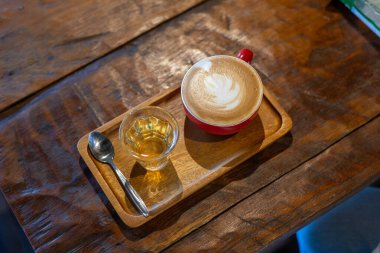 Süt köpüğüyle süslenmiş bardakta sıcak kahvenin yüksek açılı görüntüsü. Tatlıyı sevenler için temiz bir bardakta şerbet ile servis edilir. Ahşap bir tepsiye konmuş ve klasik bir görünüm için masaya servis edilmiş..