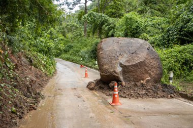Dağdan düşen büyük kayalar, yağmur mevsiminde yeşil bir ormanda kırsal beton bir yolu tıkıyor. Yola yerleştirilmiş turuncu koniler var. Tayland 'da yağışlı bir mevsim sonrasında büyük kayalar düşer ve toprak kayar..