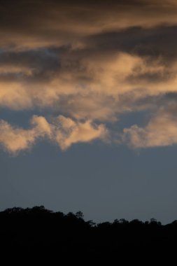 Kalın bulutların manzarası, mavi gökyüzüne vuran turuncu ışık, ağaçların, ormanların ve dağların siluetleri. Resmin ortasındaki metin için alan