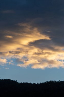 Kalın bulutların manzarası, mavi gökyüzüne vuran turuncu ışık, ağaçların, ormanların ve dağların siluetleri. Resmin ortasındaki metin için alan