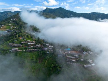 Yüksek açılı bir manzara, dağların ortasında beyaz sisle çevrili bir köy. Güzel manzaralar, ormanların ortasındaki köyler, yeşil dağlar, açık gökyüzü. Bir İHA 'dan fotoğraflar