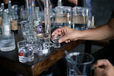 Elindeki küçük temiz bardağa ve etrafındaki cam şişelere odaklan..