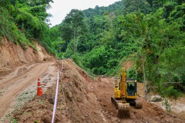 Bir kepçe, nehir kenarındaki bir yol üzerinde çalışıyor. Su yolu boyunca uzanan bir dağ ormanının içinden geçiyor. Yol yağmur mevsiminde heyelan tarafından hasar gördü.
