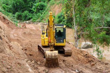 Bir kepçe, nehir kenarındaki bir yol üzerinde çalışıyor. Su yolu boyunca uzanan bir dağ ormanının içinden geçiyor. Yol yağmur mevsiminde heyelan tarafından hasar gördü.
