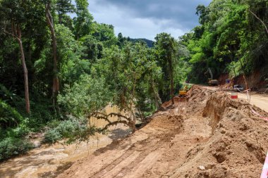 Yağmur mevsiminde, ormanın içinden geçen kırsal bir yolun peyzajı, yolun durulduğu bir kepçeyle onarılıyor. Yol Tayland 'da bir ormanda nehri takip ediyor..