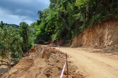 Yağmur mevsiminde, ormanın içinden geçen kırsal bir yolun peyzajı, yolun durulduğu bir kepçeyle onarılıyor. Yol Tayland 'da bir ormanda nehri takip ediyor..