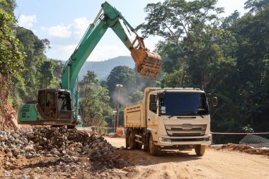 Beyaz bir kamyon yeşil bir kepçenin çevresi ormanla çevrili bir şantiyenin arkasına kum yüklemesini bekliyor..