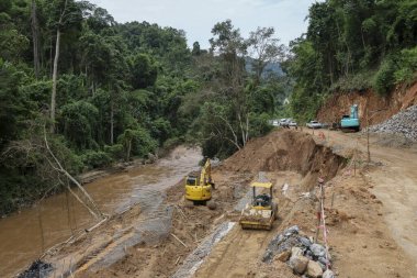 Kırsal yolun manzarası bir yol müteahhiti tarafından bir kepçe tarafından onarılıyor. Yol, yağmur mevsiminde nehir boyunca uzanan heyelan boyunca ormanın içinden geçiyor..