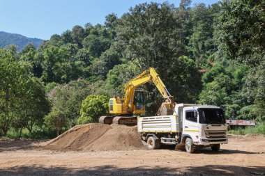 Beyaz bir kamyon, çevresi ormanla çevrili bir şantiyenin arkasına kum yüklemek için sarı bir kepçeyi bekliyor..