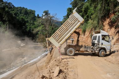Beyaz bir kamyon, bir yol onarım alanının inşaat alanında tamir edilen bir ormanın içinden geçen kırsal bir yola kum döküyor. 