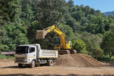 Beyaz bir kamyon, çevresi ormanla çevrili bir şantiyenin arkasına kum yüklemek için sarı bir kepçeyi bekliyor..