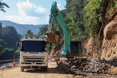 Beyaz bir kamyon yeşil bir kepçenin çevresi ormanla çevrili bir şantiyenin arkasına kum yüklemesini bekliyor..