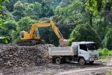 Bir kepçe, yol bariyeri yapmak için büyük bir kaya yığınını beyaz bir kamyona yüklüyor..