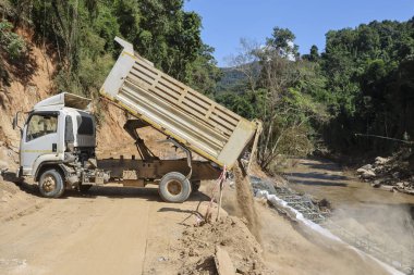 Büyük bir çöp kamyonu verimli yeşil dağlar ve ağaçlarla çevrili engebeli toprak yola ağır kahverengi toprak yükünü boşaltıyor. Güçlü araç, altyapı iyileştirmesi üzerinde gayretle çalışıyor