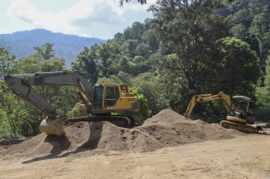 Ağır ekipmanlar inşaat alanını işletiyor, kazıcıyla büyük toprak yığınlarını hareket ettiriyor. Güçlü makineler, yeşil dağların ve sık ormanların zeminine karşı toprak kaldırma çalışmaları gerçekleştiriyor