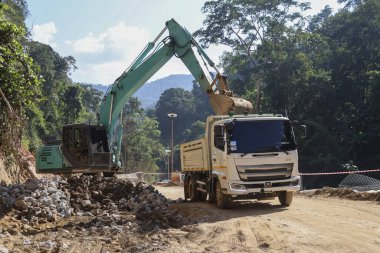 Ağır kazıcı, toprak hareket ettirme çalışmalarını zorlu bir inşaat alanında gerçekleştiriyor. Güçlü makineler çöp kamyonuna malzeme yükler. Yoğun dağ ormanlarında yeni bir yol yaratır.