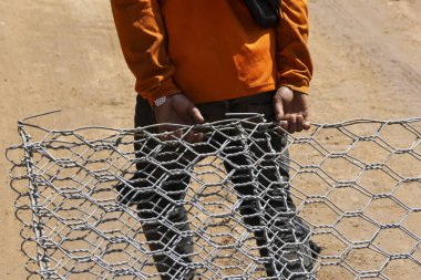 İşçi sağlam metal tel örgüleri kavrıyor ve ağır işçilik sırasında özenli bir şekilde odaklanıyor. Turuncu uzun kollu bluz, koyu renk pantolon. Açık hava inşaat sahnesi, kumlu toprak görünür, kararlı çaba