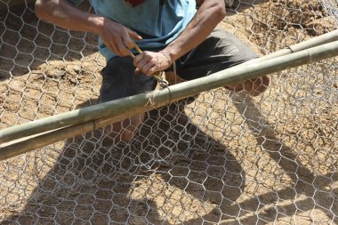 Çalışkan işçi, odaklanmış elleriyle bambu direğine tel örgü örmek için pense kullanıyor. Bir açık hava inşaat projesi malzemelerin bağlanmasıyla ilerliyor. Çalışkan bireyler gösteriler