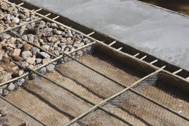 Çakıl taşlarının üzerinde demir destek ızgarası olan yeni beton zemin çalışması. İnşaat süreci ağ materyali ve hazırlık tabanı gösteriyor. İnşaat alanında sürekli ilerleme kaydedildiğini gösteriyor.