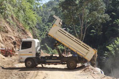 Beyaz çöp kamyonu özenle çalışıyor, yatağını kaldırıyor ve toprağı engebeli toprak yola bırakıyor. Sahne, doğal dağlara meydan okuyan belirlenmiş çaba inşaatı ve altyapı gelişimini yakalıyor