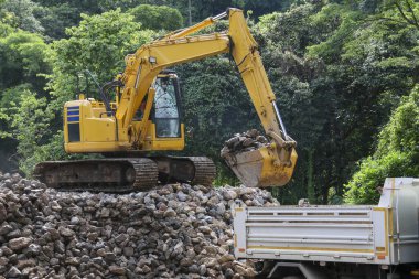 Sarı kazıcı, güçlü ağır ekipman, kaya malzemelerini büyük çöp kamyonuna yüklüyor. Temel endüstriyel çalışma açık hava inşaat alanı, altyapı geliştirme projeleri