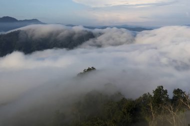 Yumuşak bulutlarla dolu puslu dağ manzarası yemyeşil tepeler üzerinde yuvarlanarak doğal güzellik ve sükunetin huzurlu atmosferini yaratıyor.