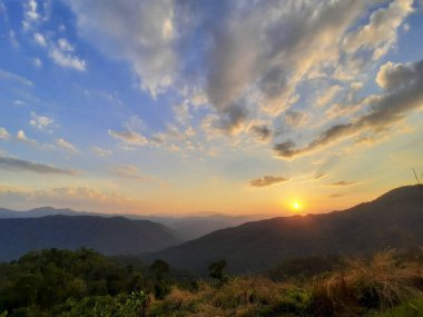 Yuvarlanan tepelerde gün batımı, sıcak renklerle boyanmış, huzur ve merak hissi uyandırıyor. Gökyüzü turuncu ve sarı ışıkla parıldıyor