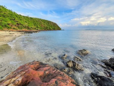 Huzurlu okyanus ve yemyeşil yamaçlı sakin kıyı manzarası. Berrak mavi su yavaşça kıyıya vurur, çakıl ve kayaları ortaya çıkarır. Bu huzurlu sahne, sakin ve doğal güzelliği çağrıştırıyor.