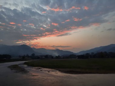 Dağların ve tarlaların üzerinde, yumuşak pembe ve turuncu renklerle boyanmış huzurlu bir günbatımı. Dolambaçlı yollar huzur ve sükuneti çağrıştıran huzurlu manzaraya götürür.