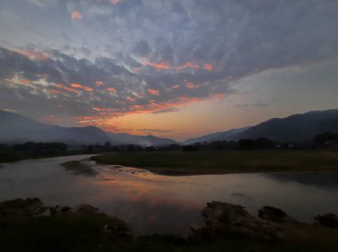 Görkemli günbatımı gökyüzünü canlı renklerle boyar, dingin nehir ve yuvarlanan tepeler üzerine sıcak bir parıltı saçar, huzur ve doğal güzelliği çağrıştırır.