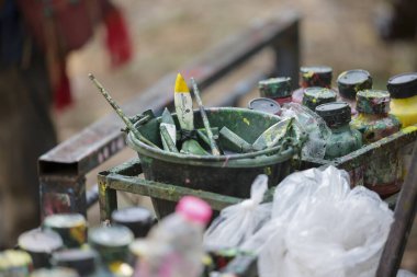 Yaratıcı ve dağınık sanatçı tezgahı malzeme dolu. Stüdyo ortamında renkli boyalarla çevrili yeşil kovada kullanılan fırça ve aletler sanatsal kaosu gösteriyor.
